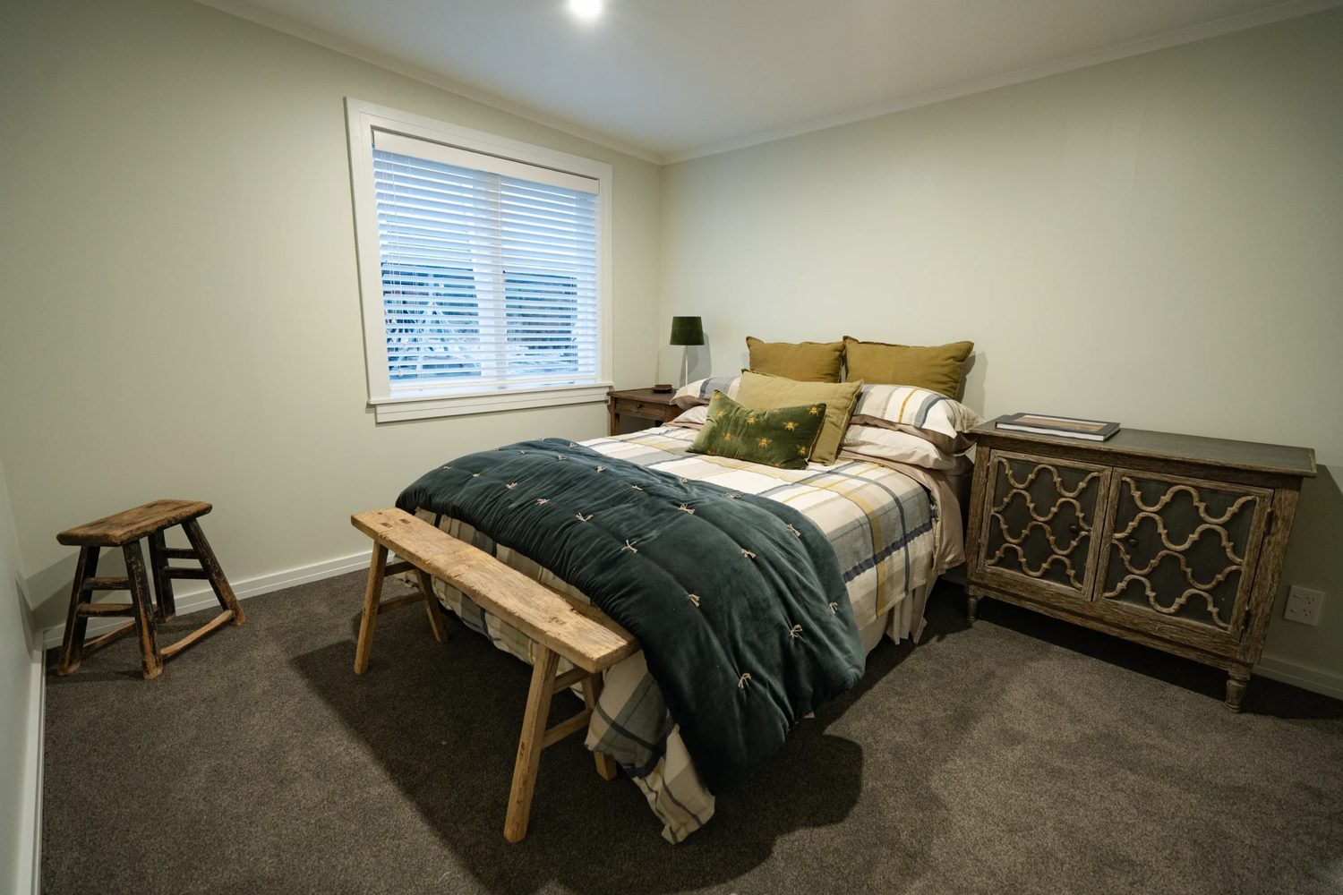 Bedroom with grey carpet, a made bed with a dark green throw and a carved timber sideboard