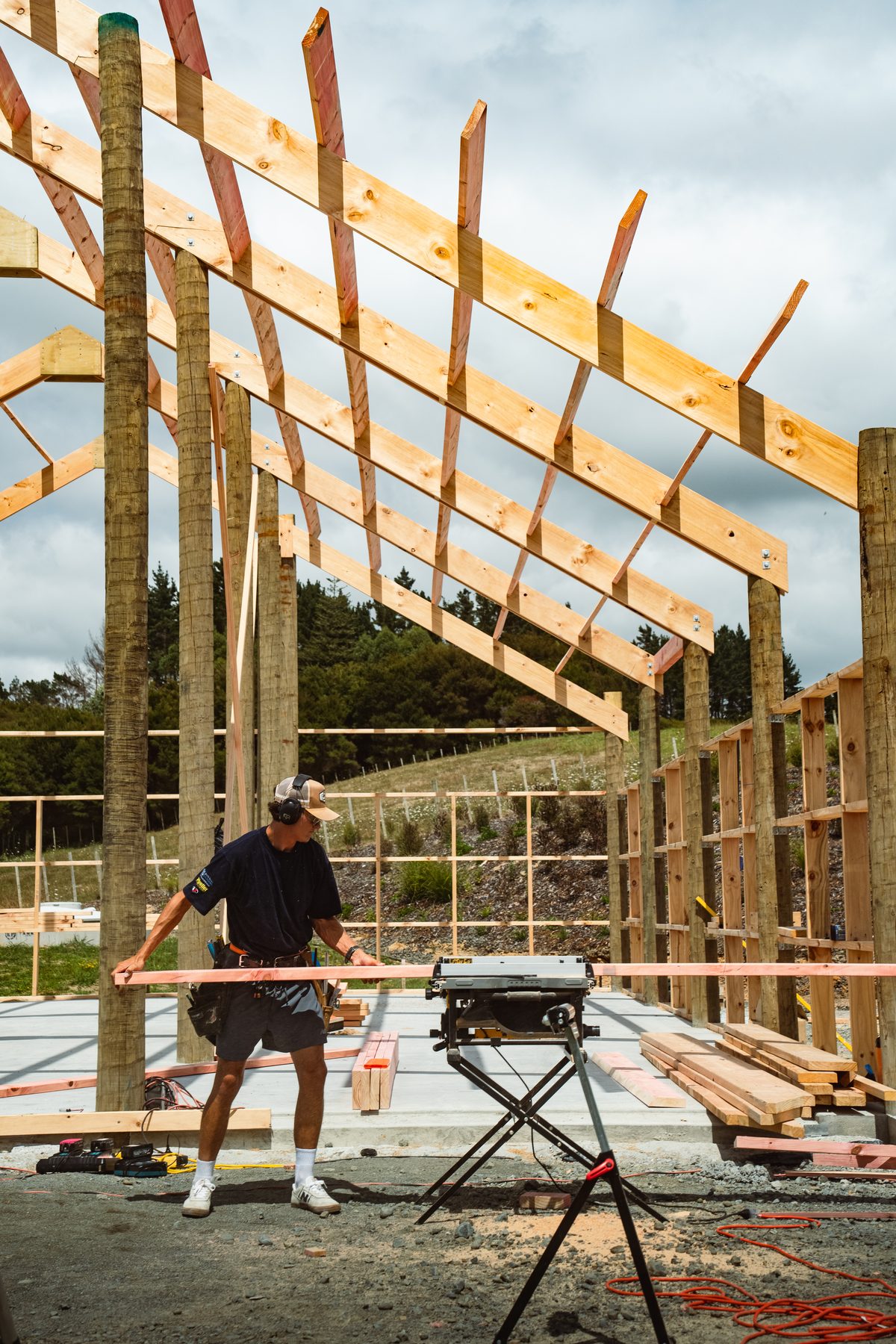 Builder cutting timber at a saw bench on the slab inside a pole shed frame