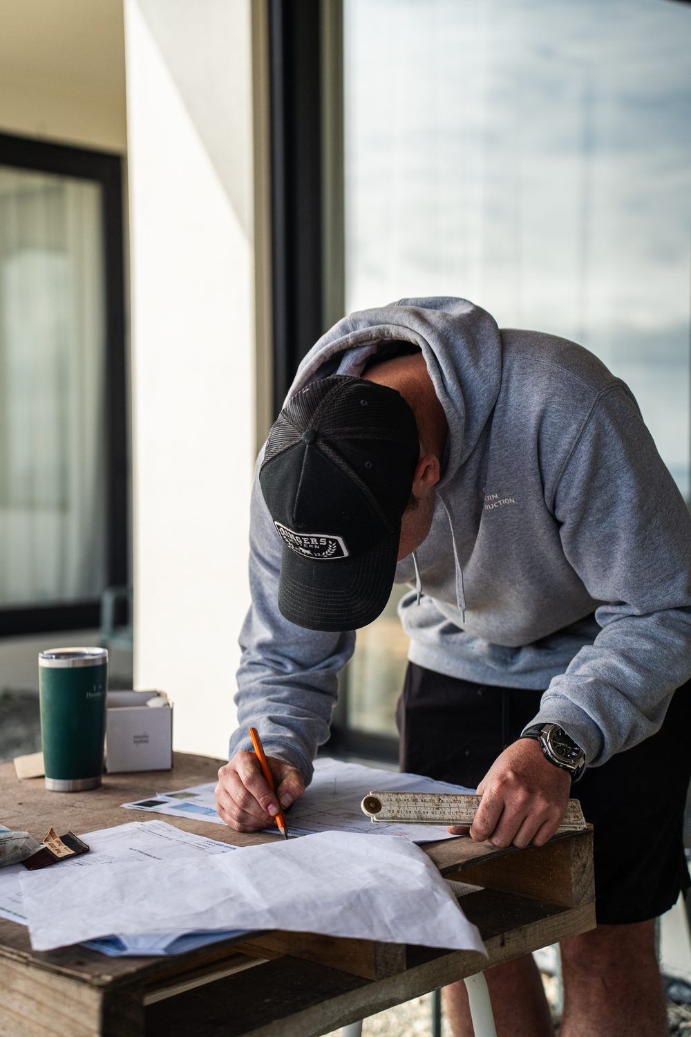 Builder leaning over a set of plans with a pencil, a drink bottle beside him