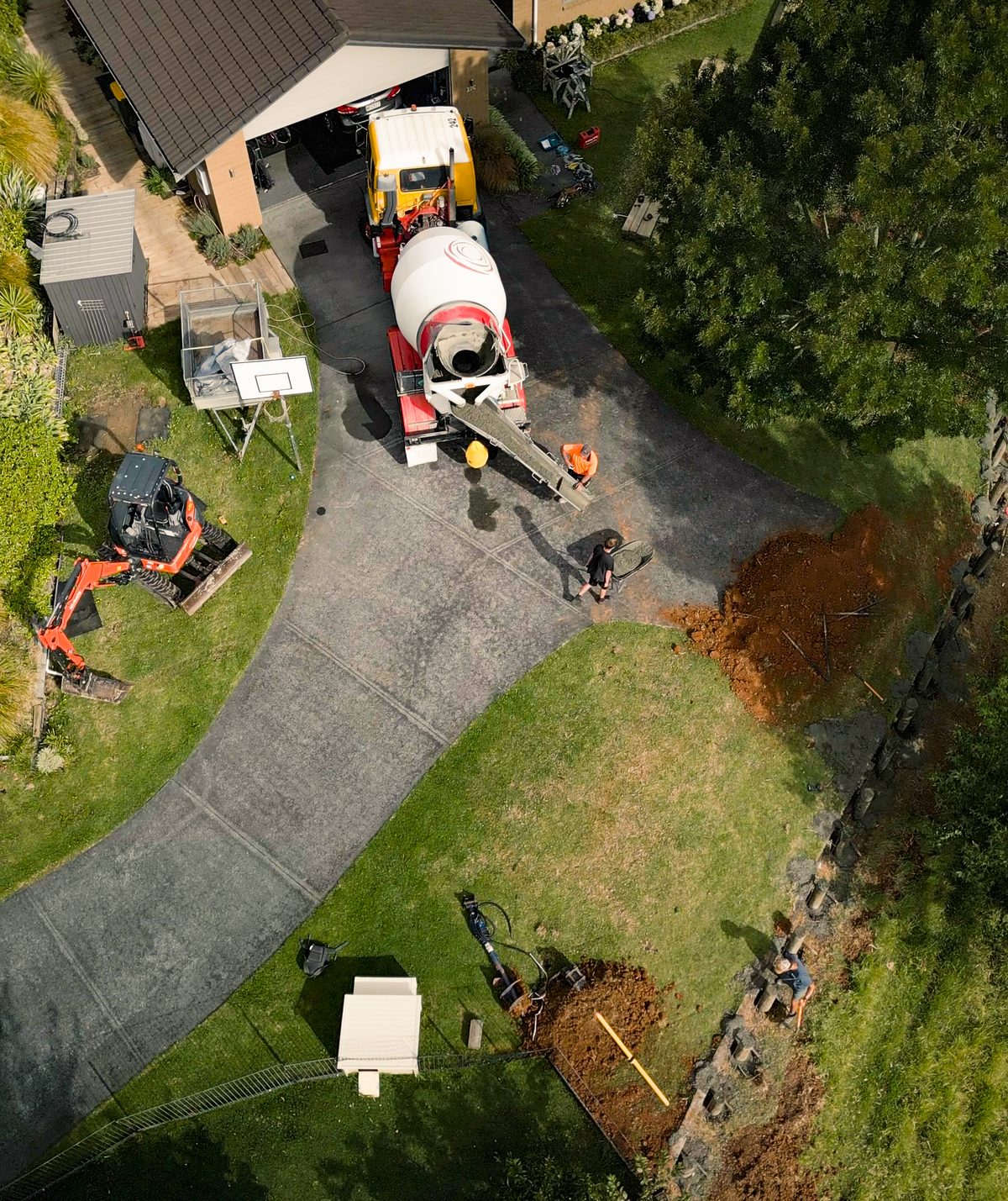 Overhead view of a concrete truck on a driveway, a mini excavator and a trench dug along a boundary