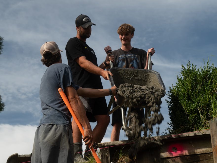 Three workers tipping concrete from a wheelbarrow into a post hole behind a timber retaining wall