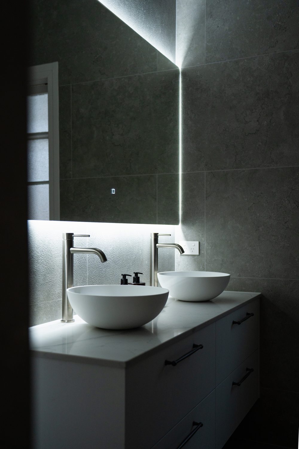 Two vessel basins on a vanity lit by a backlit mirror in a dark tiled bathroom
