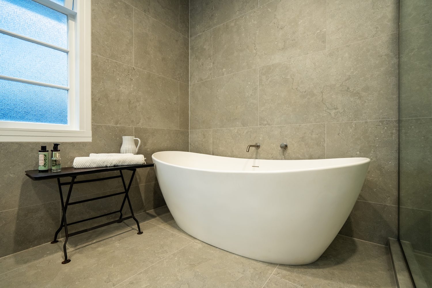Freestanding white bath against grey tiled walls, a small table with towels beside it