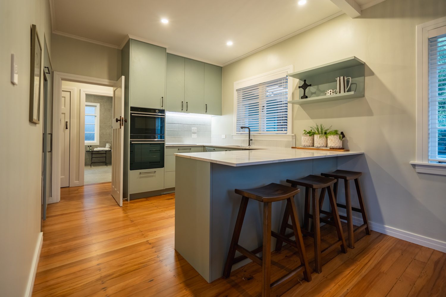 Kitchen with sage green cabinetry, wall ovens and a breakfast bar with three timber stools