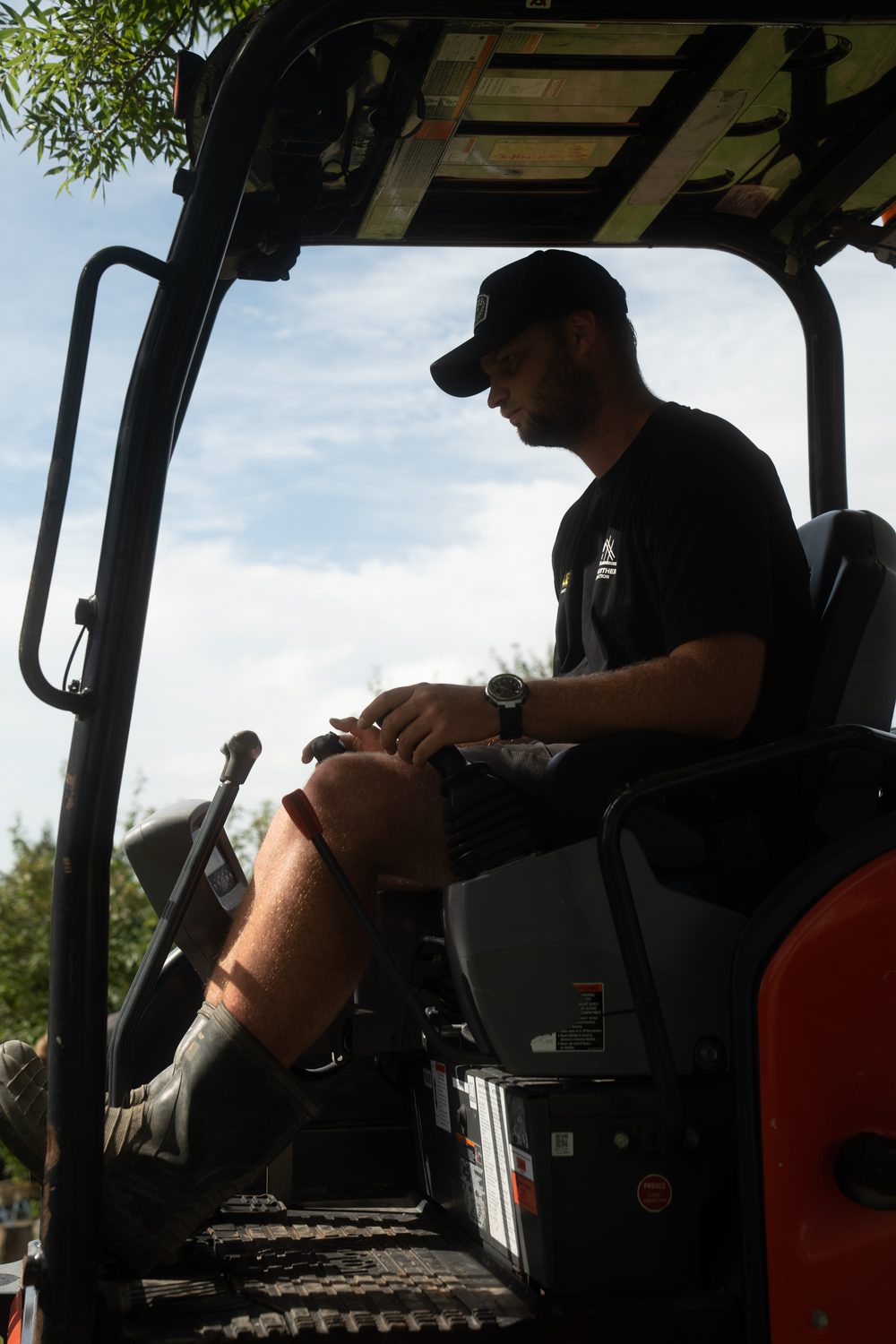 Machine operator in a cap seated in a mini excavator, silhouetted against the sky