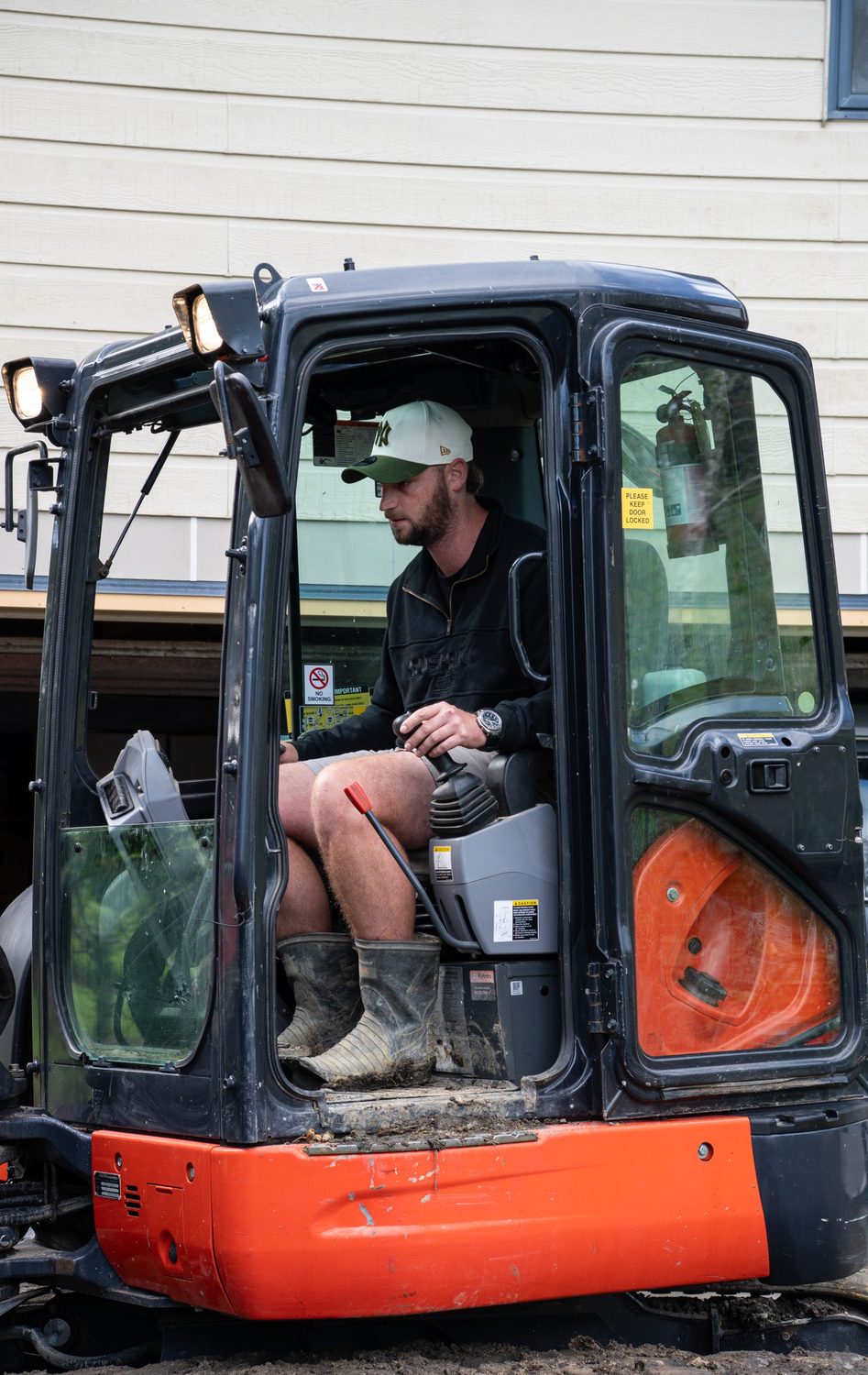 Operator in a cap and work boots sitting in the cab of an orange mini excavator
