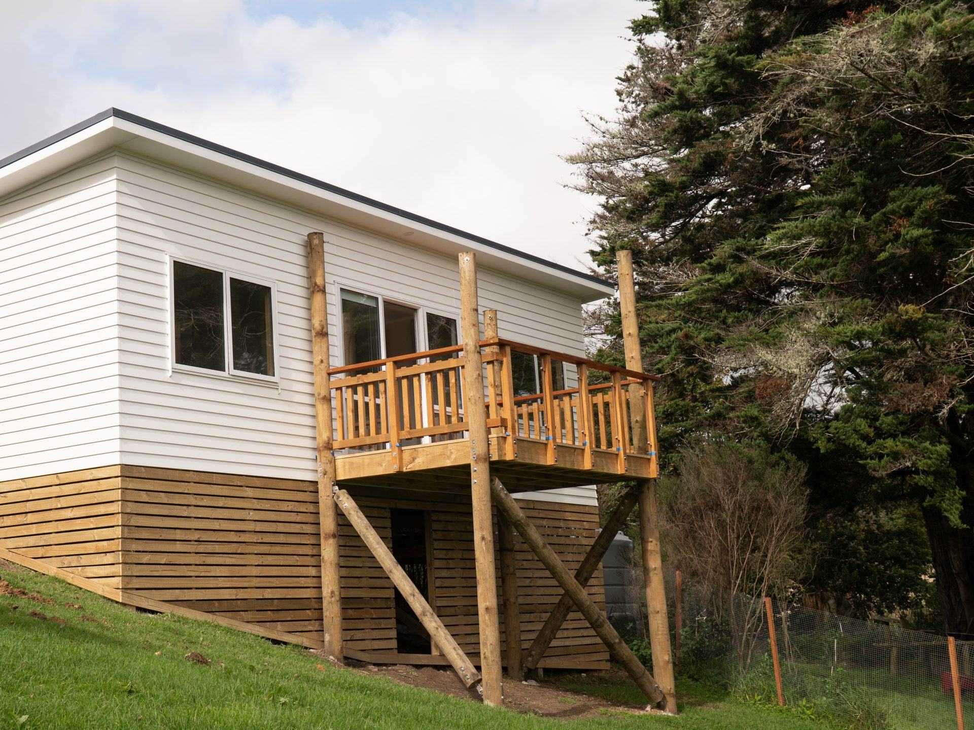 White weatherboard building on a slope with a raised timber deck and balustrade held up on braced poles