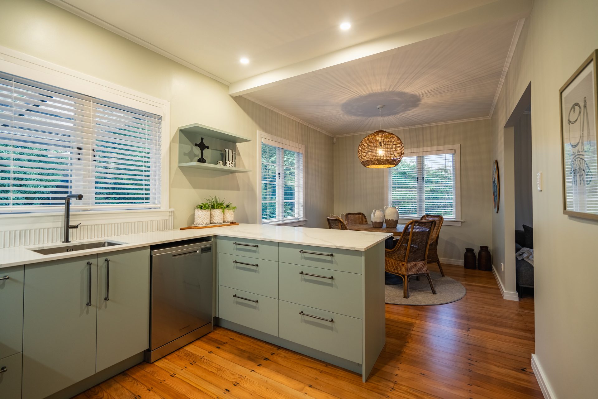 Renovated kitchen with sage green cabinets and a stone benchtop, opening to a dining area under a woven pendant