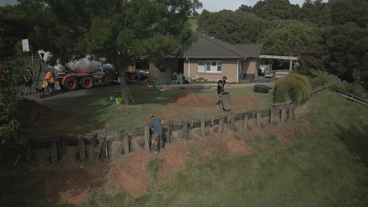 Crew at work on a timber retaining wall across a sloping lawn, concrete truck on the driveway