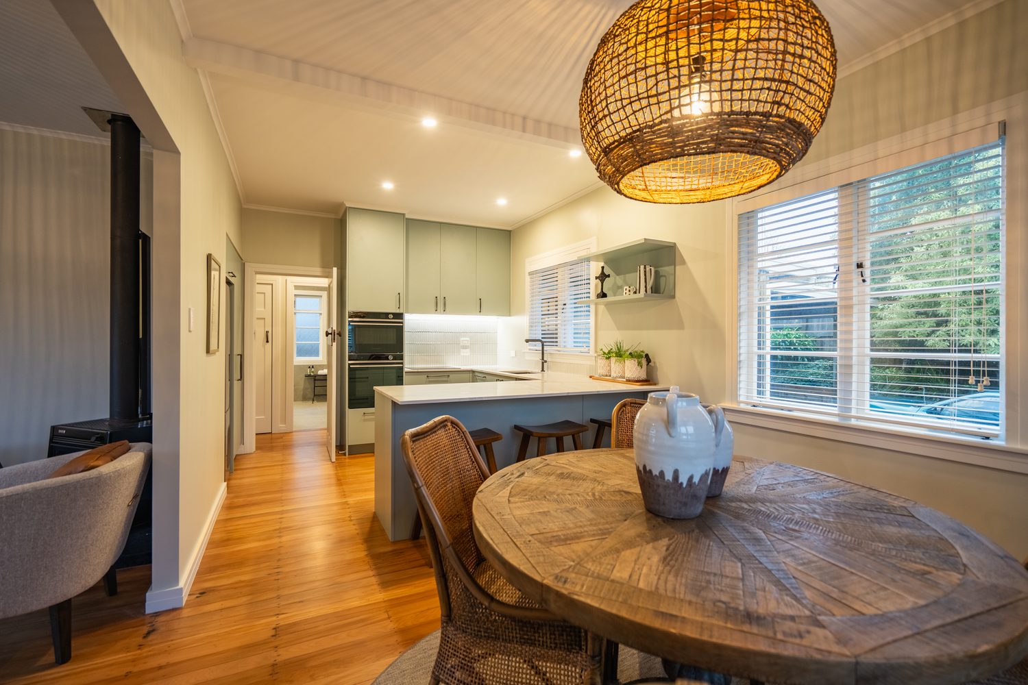 Round timber dining table under a woven pendant, the kitchen and breakfast bar behind