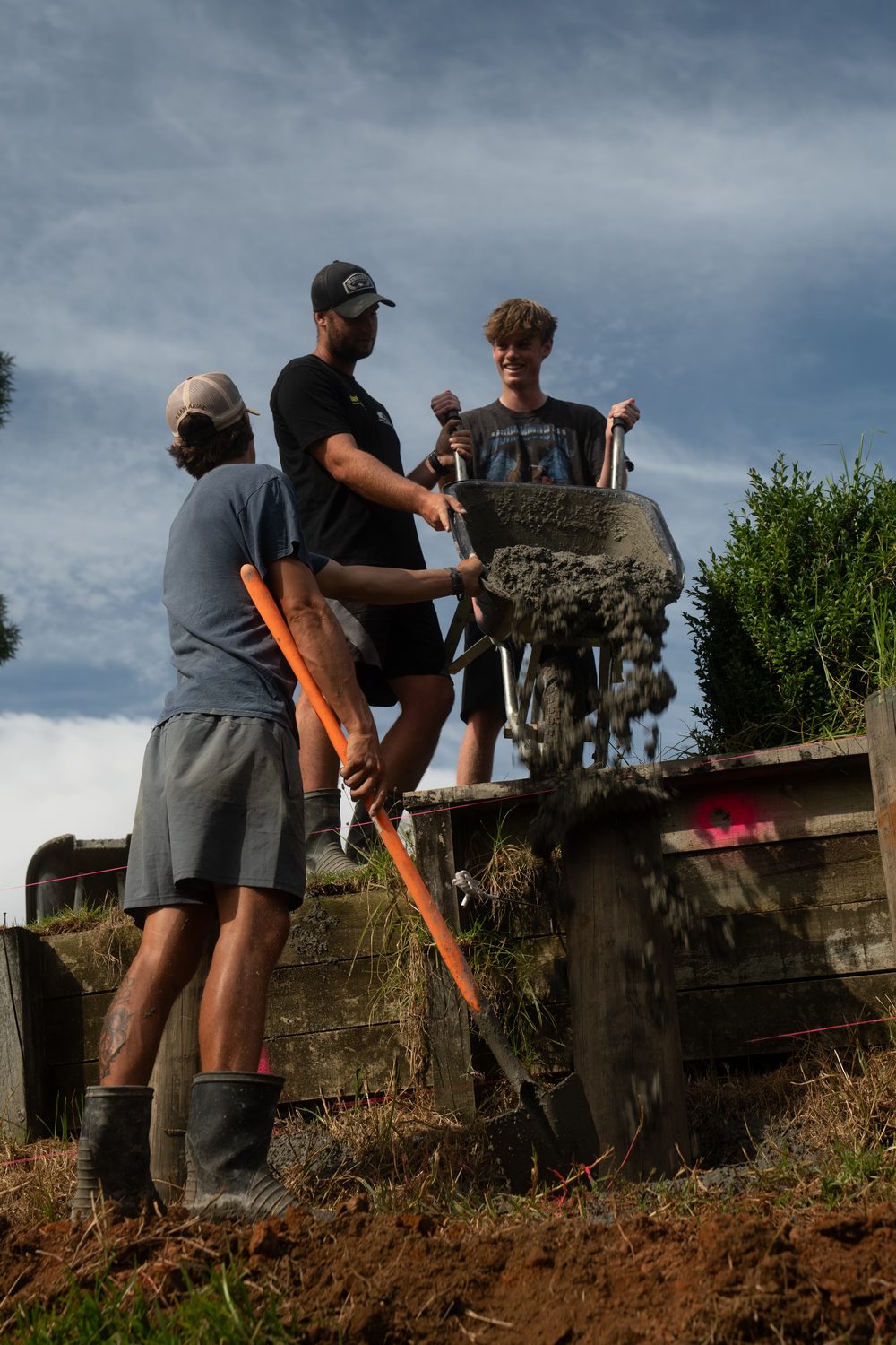 Workers shovel concrete from a wheelbarrow into a hole behind a timber retaining wall