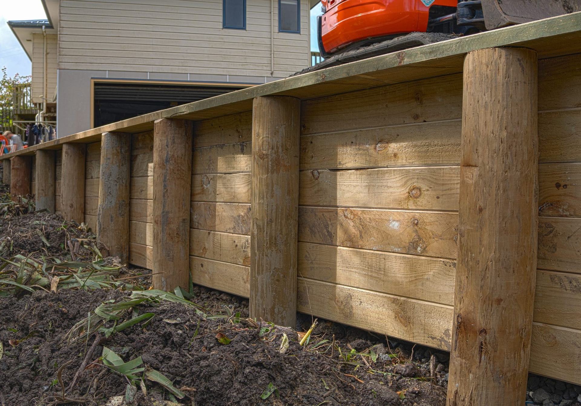 Timber retaining wall of round poles and horizontal boards, with a mini excavator on the level ground above